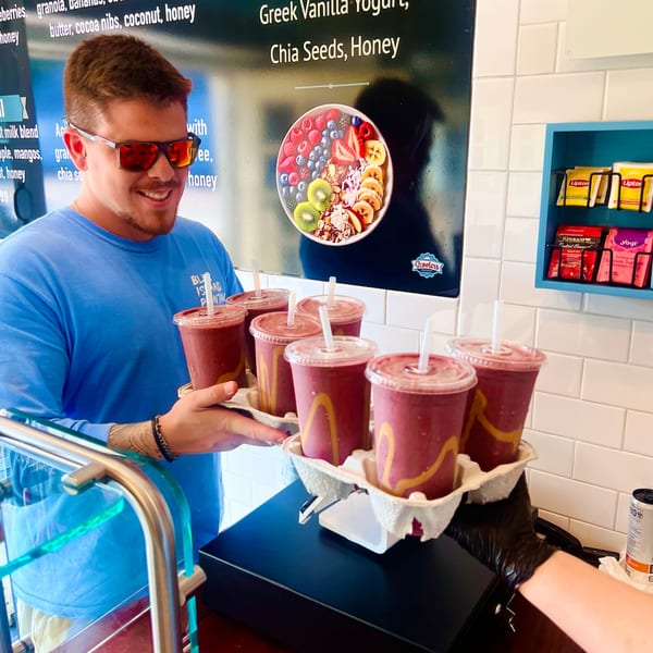 A Scooters employee in sunglasses hands over two carriers of smoothies to a customer.