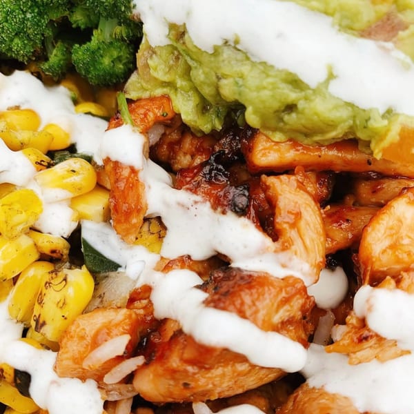 Close-up of a healthy bowl with chicken, broccoli, corn, guacamole, and a creamy white sauce.