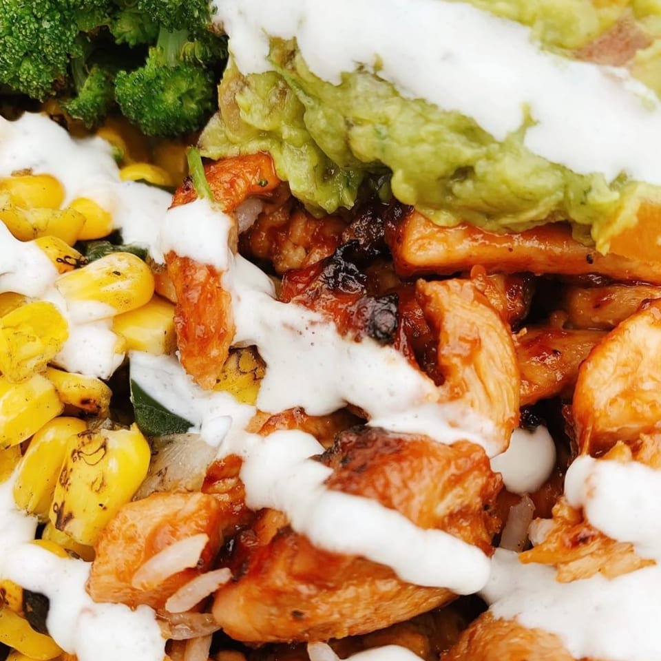 Close-up of a healthy bowl with chicken, broccoli, corn, guacamole, and a creamy white sauce.