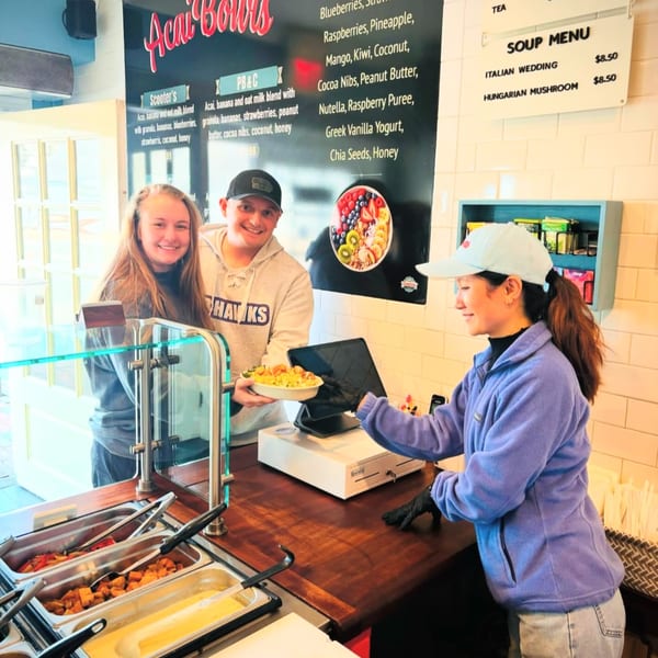 Scooters' employee serves a healthy acai bowl to two smiling customers at the counter.