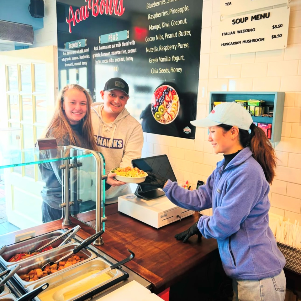 Scooters' employee serves a healthy acai bowl to two smiling customers at the counter.
