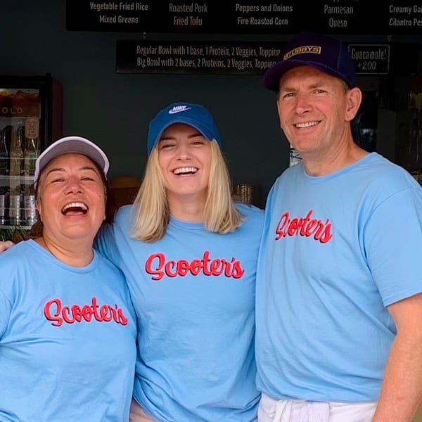Three smiling Scooters employees in light blue shirts and hats stand together, with a menu board behind them.