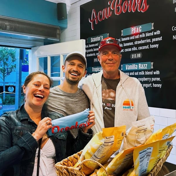 Three smiling people, two men and one woman, stand in front of a Scooter's Açaí Bowls menu.