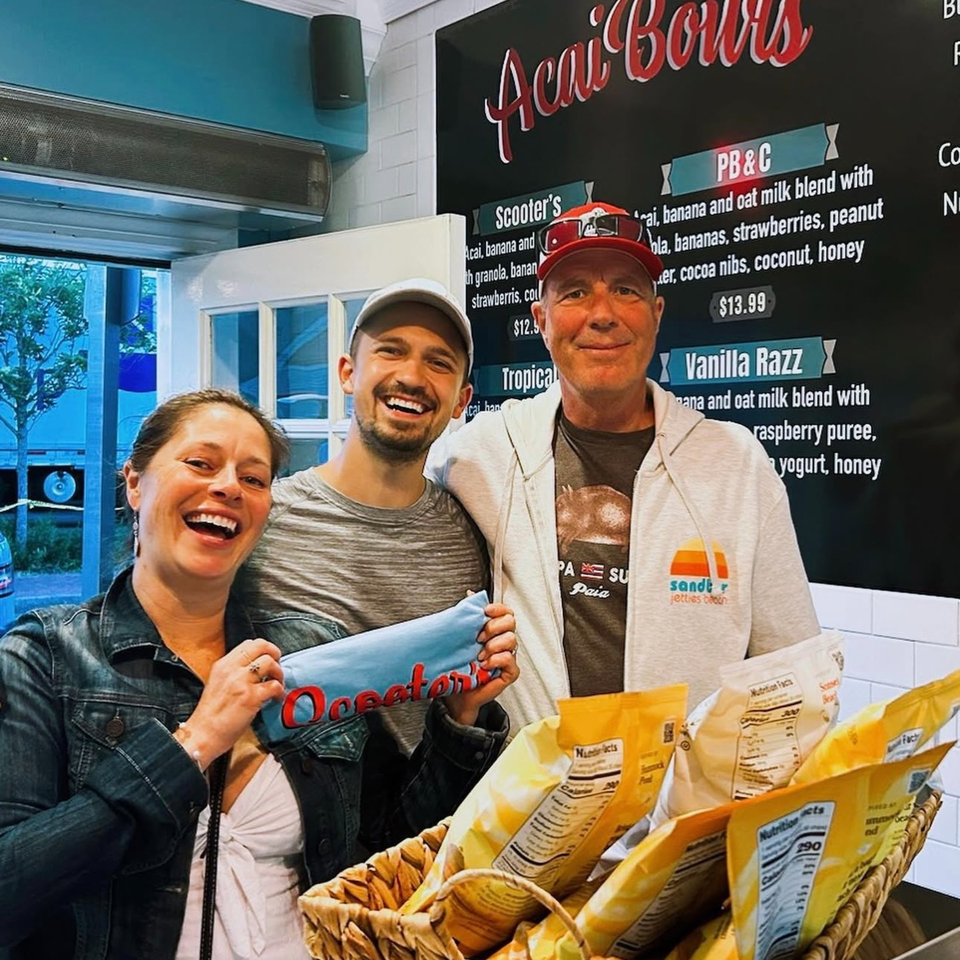 Three smiling people, two men and one woman, stand in front of a Scooter's Açaí Bowls menu.