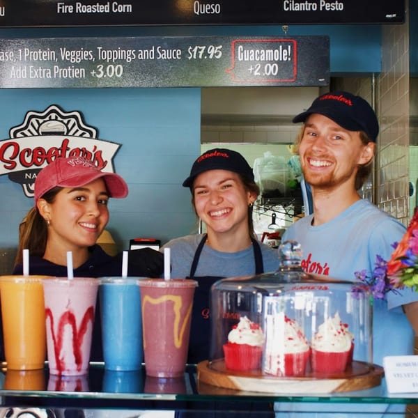Three smiling Scooters employees stand behind a counter with colorful smoothies and red velvet cupcakes.