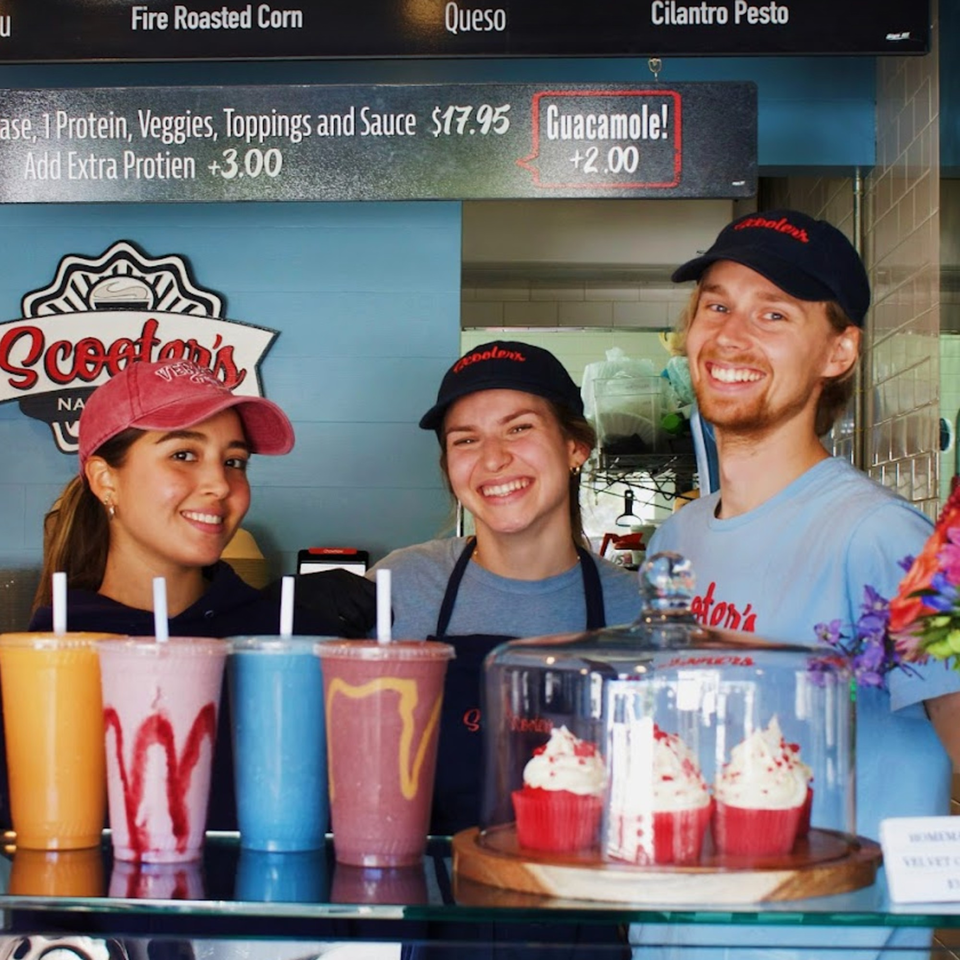 Three smiling Scooters employees stand behind a counter with colorful smoothies and red velvet cupcakes.