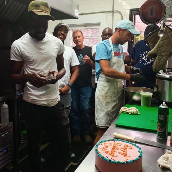 A group of men in a kitchen, one wearing an apron, with a birthday cake on the counter.