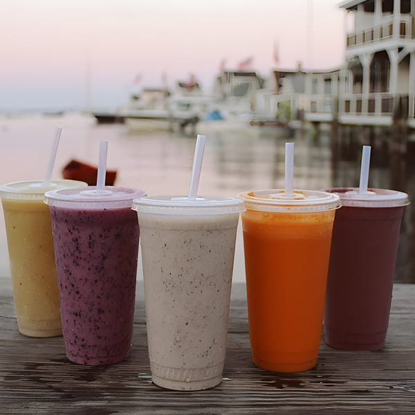 Five colorful Scooters smoothies and juices with straws on a wooden deck overlooking a harbor.
