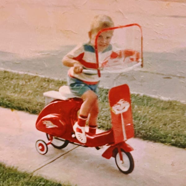 A young child in a striped shirt and shorts rides a red toy scooter on a sidewalk.