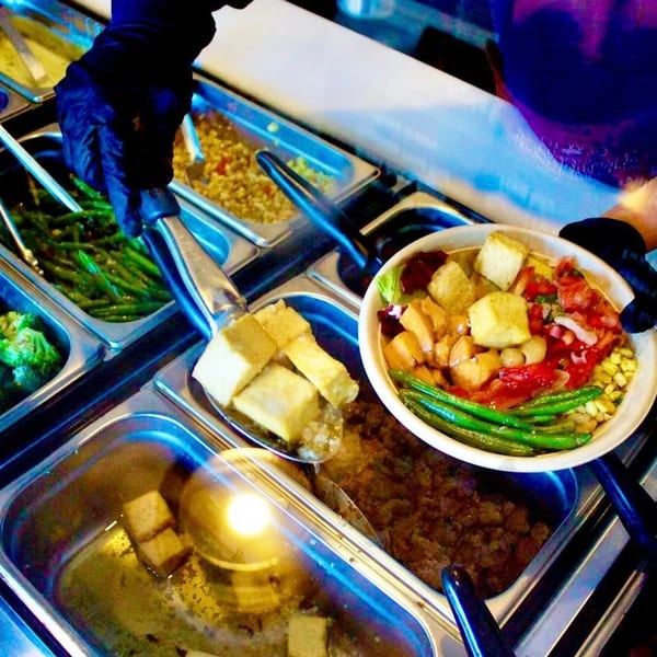 A Scooters employee in black gloves serves healthy food from a buffet line into a bowl.